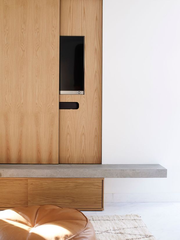 A minimalist interior detail featuring a full-height light timber wall and integrated black fireplace or media panel with a slim horizontal control or storage element. A grey concrete ledge projects above matching timber cabinetry, while a white wall, pale floor, textured rug, and partial tan leather seating create a clean, warm contemporary composition.
