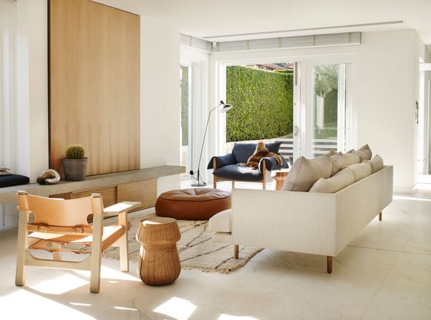 Bright, refined contemporary living room with white walls and pale flooring, a large cream sectional sofa, navy lounge chair, leather butterfly chair, and sculptural timber and leather furnishings. Full-height glazed doors and windows open toward a leafy courtyard, while a warm timber feature panel and low built-in joinery anchor the room. Natural light, neutral tones, and organic materials create an airy, calm atmosphere.