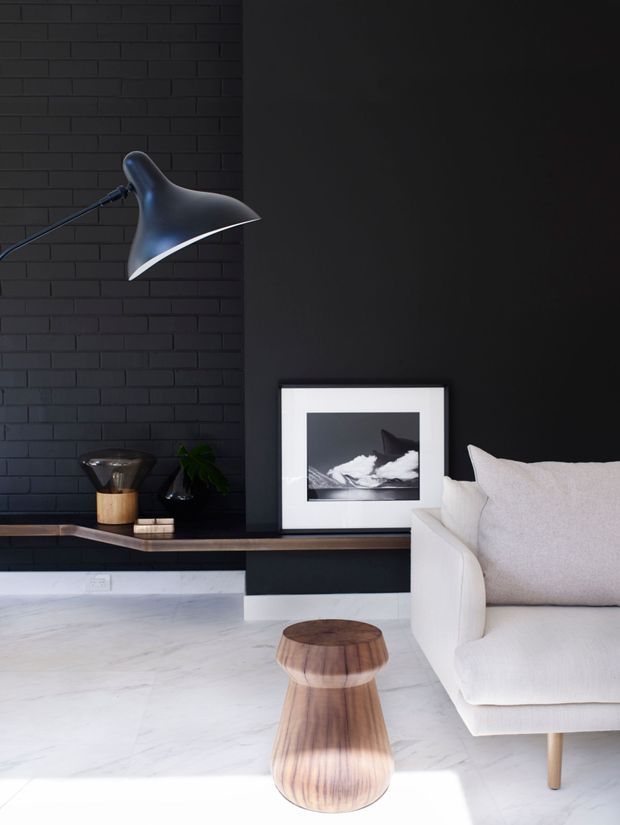 A refined contemporary living room vignette with a dramatic black brick feature wall and adjacent matte black wall, a sculptural black wall lamp, floating dark timber and black joinery, framed monochrome artwork, and a white upholstered armchair. Pale marble-look flooring and a sculptural timber side table provide contrast, creating a high-contrast, minimal interior.