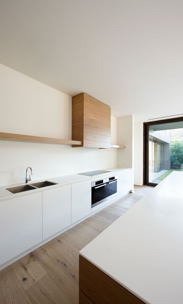 A bright, minimalist contemporary kitchen with a long white run of handleless cabinetry, integrated black oven, induction cooktop, stainless-steel double sink and slim timber open shelving. A timber-clad rangehood enclosure provides the main feature, while pale timber flooring and a large white island create a clean, warm scheme. Full-height glazed doors connect the kitchen to a small landscaped courtyard.