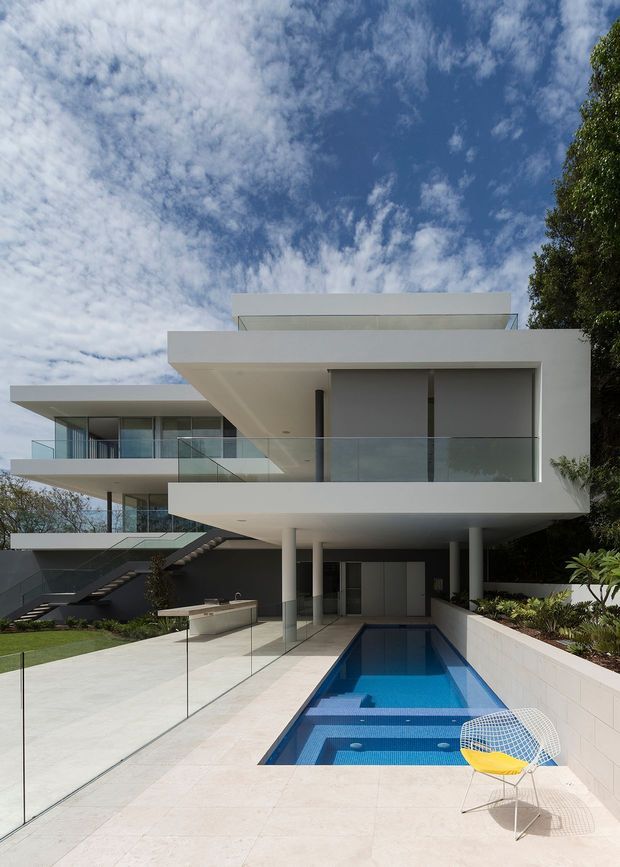 Exterior view of a large modern multi-level residence with crisp white cantilevered volumes, extensive glass balustrades, floor-to-ceiling glazing, and a flat roof. A narrow rectangular swimming pool runs alongside a pale paved terrace, with an outdoor kitchen or bench area and landscaped planting. An external stair connects the upper and lower levels, and mature trees frame the property.