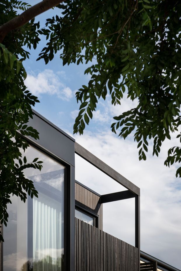 Vertical architectural photograph of a contemporary house exterior framed by leafy tree branches. The building features crisp rectangular volumes, dark metal or painted frames, expansive glazing, vertical timber screening, and a projecting open rectangular structure against a partly cloudy sky.
