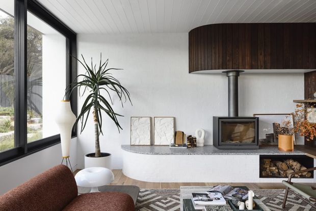A refined mid-century-inspired living room centred on a sculptural curved fireplace hearth with a dark timber mantel and black wood-burning stove. Floor-to-ceiling black-framed glazing opens to greenery, while a white painted timber ceiling, textured white walls, terrazzo-like hearth surface, patterned rug, sculptural furnishings, and a tall indoor plant create a warm, sophisticated interior.