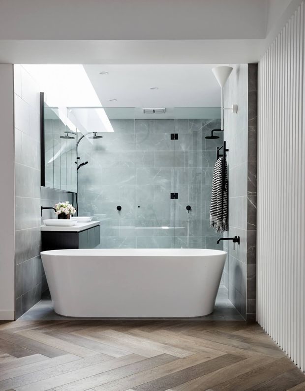 Bright contemporary bathroom featuring a freestanding white bathtub, glass-enclosed walk-in shower, wall-mounted black fixtures, grey stone-look wall tiles, a dark floating vanity with vessel basins, and pale timber flooring laid in a herringbone pattern. A vertical white ribbed wall treatment adds texture along the right side.