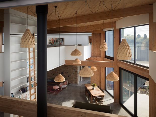 A warm, double-height open-plan interior viewed from an upper-level landing or mezzanine. The space features extensive timber ceiling and wall lining, exposed dark brickwork, white built-in shelving and cabinetry, large black-framed windows, and numerous woven pendant lights suspended at different heights. A dining table, seating area, and lower-level living zone are visible, along with a black cylindrical flue or structural column.