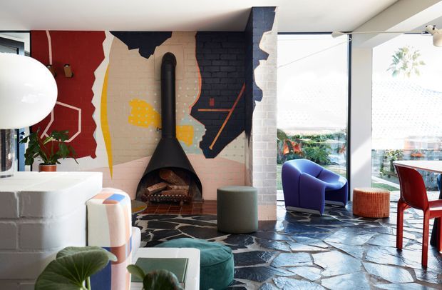 Eclectic, colourful living room with a prominent black sculptural fireplace and exposed brick chimney breast, graphic mural artwork, irregular dark stone flooring, and large glazed openings. Curved white built-in seating and contemporary upholstered furniture create a playful, art-led interior, with a dining area visible to the right.