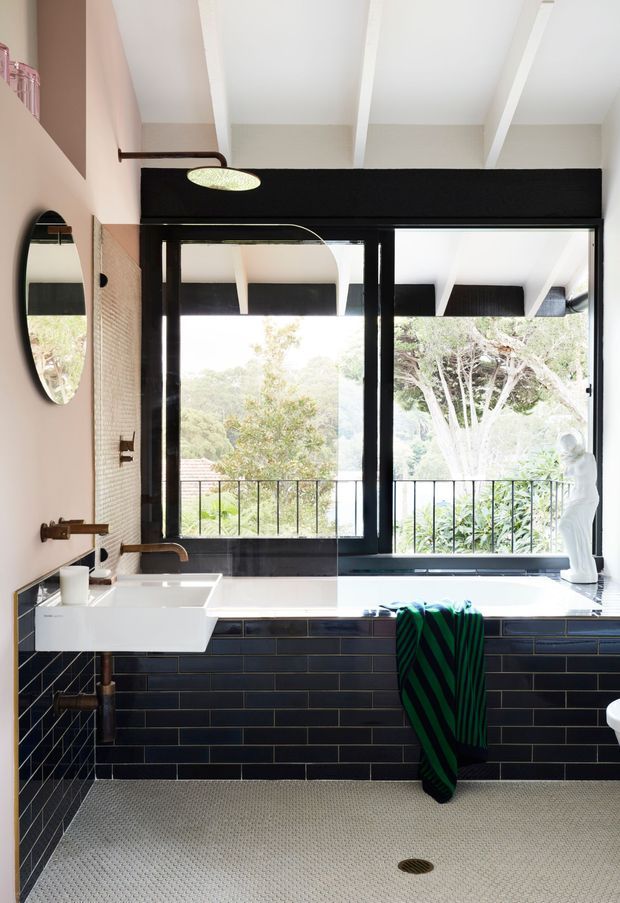 A bright, design-focused bathroom with a freestanding white vessel sink, exposed brass plumbing, black-framed windows and a large white tub or ledge beneath them. The walls and tub surround feature glossy dark navy rectangular tiles with light grout, while the floor is covered in small light-grey mosaic tiles. A pink-toned wall, oval mirror, wall-mounted light, and green-and-black towel add colour. The windows open to leafy vegetation and a balcony railing outside, and the ceiling has exposed white beams.