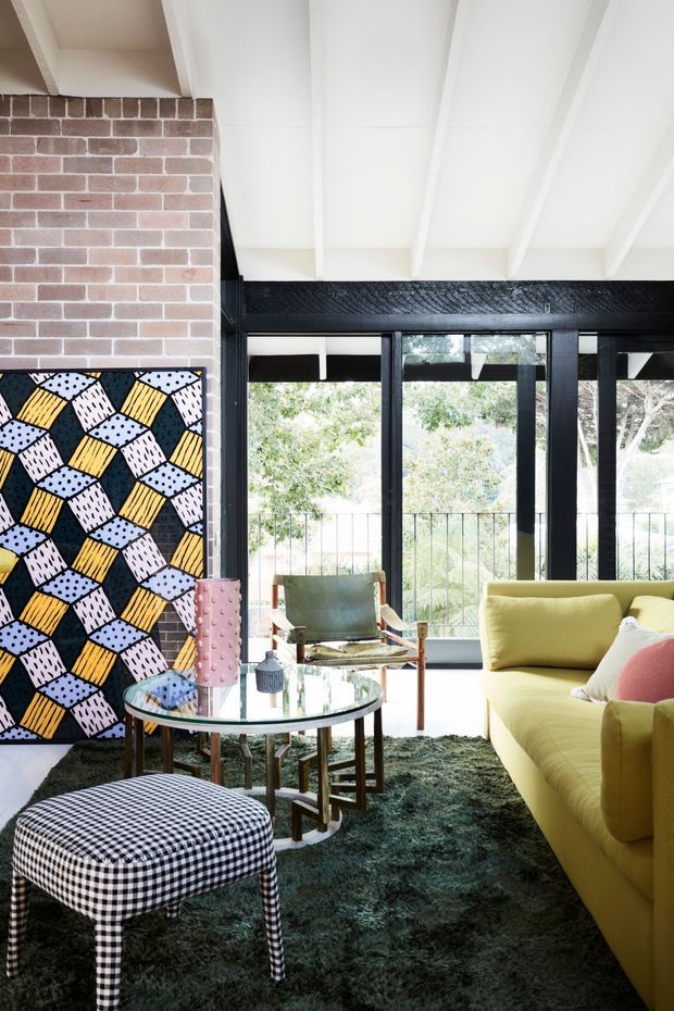 A lively living room with a white exposed-beam ceiling, exposed red brick wall, black-framed floor-to-ceiling glazing, and views to greenery outside. The room features a mustard-yellow sofa, vintage-style green armchair, round glass coffee table, patterned black-and-white ottoman, colourful geometric artwork, and a dark textured rug. The overall look is playful, eclectic, and retro-inspired.