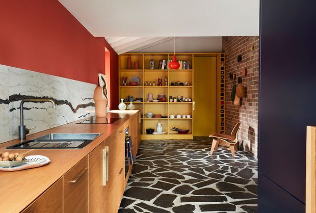 Bold, richly coloured kitchen with warm timber cabinetry and island bench, a dramatic white veined stone splashback, open ochre shelving, and a distinctive irregular dark stone floor. A red pendant and red wall contrast with an exposed brick feature wall, while a small sitting area is visible at the far end.