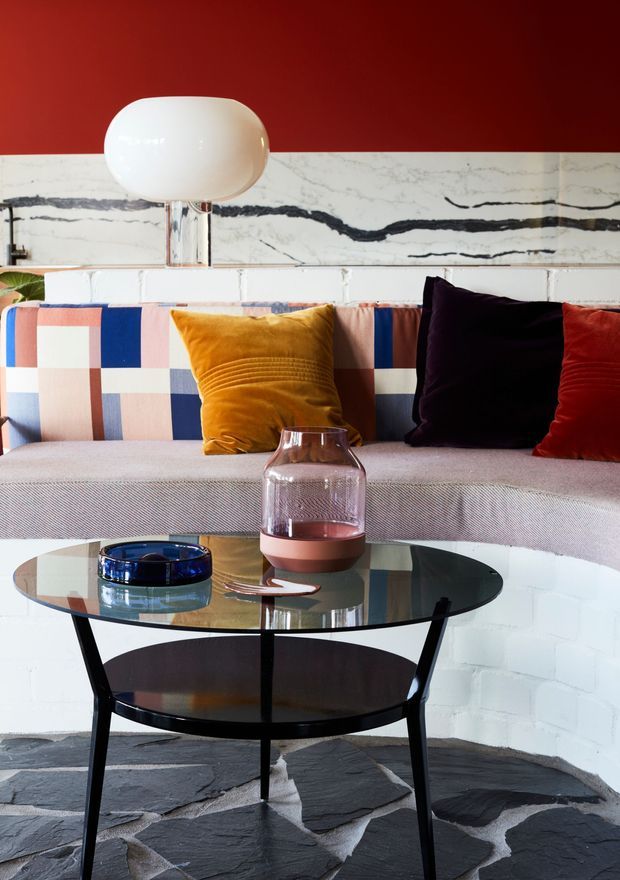 A vividly styled living room vignette featuring a curved pale sofa with a geometric multicolour upholstered back and cushions in mustard, navy, burgundy and red. A round glass-topped black coffee table sits in front, with decorative vessels and a blue ashtray. The room has a dramatic red painted upper wall, a white marble-look horizontal feature surface with dark veining, white painted brickwork, and an irregular dark slate floor. A sculptural white globe table lamp is positioned behind the sofa.