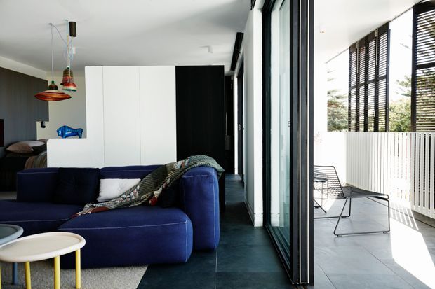 A contemporary open-plan living area with a deep navy sectional sofa, dark tiled flooring, and a compact bedroom zone partially screened by black-and-white built-in cabinetry. Full-height black-framed glazing opens onto a bright tiled terrace with a white slatted privacy screen and dark horizontal louvres. Colourful pendant lights and patterned textiles add eclectic accents to the otherwise monochromatic interior.