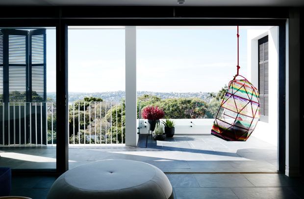 Bright contemporary indoor-outdoor living area opening onto a balcony or terrace, with expansive black-framed sliding glass doors, pale tiled flooring, white structural columns and railings, exterior louvred shutters, potted plants, and a colourful suspended hanging chair. A leafy suburban landscape is visible beyond the balustrade.
