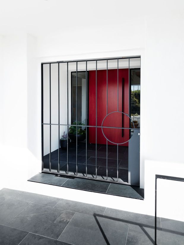 A minimalist exterior entry viewed from a paved terrace, featuring a black metal security gate with vertical bars and a circular geometric detail. Behind it is a bold red pivot door set within a dark tiled entry vestibule, framed by crisp white walls. Strong sunlight creates sharp shadows across the dark stone paving and facade.