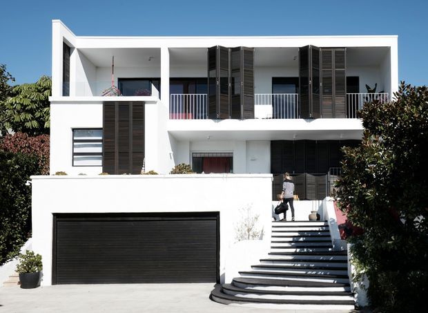 Exterior view of a contemporary multi-storey white-rendered residence with a flat roof, strong geometric forms, dark louvred screens, balconies with metal balustrades, a broad black garage door, and a stepped entry stair. Landscaping partially frames the facade.