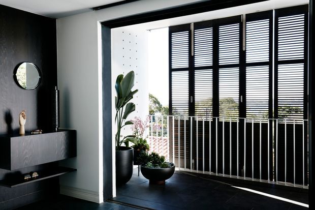 A refined contemporary interior featuring a dark built-in media console against a charcoal wall, a large opening with black-framed louvred shutters, and a balcony railing beyond. Potted plants soften the monochromatic scheme, while strong daylight filters through the shutters onto the dark floor. The image conveys a sleek, high-contrast living space with indoor-outdoor connection.