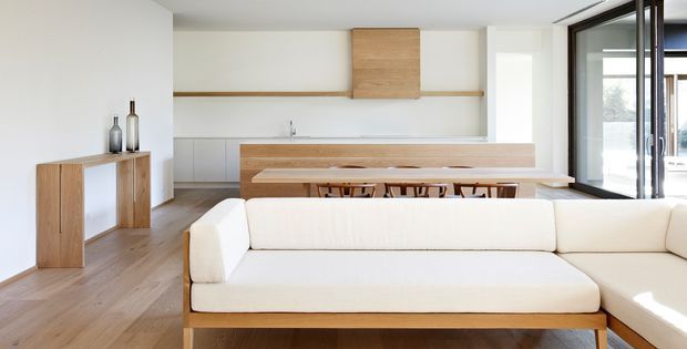 Bright open-plan living and dining area with a minimalist linear kitchen in the background. The room features pale timber flooring, white walls and cabinetry, a long timber dining table with stools, a low timber sideboard, and a large cream sectional sofa in the foreground. Full-height dark-framed glazing brings in abundant natural light.