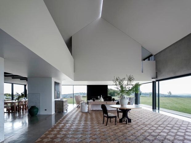A spacious, light-filled contemporary interior with a dramatic angular, vaulted ceiling and extensive floor-to-ceiling glazing overlooking open countryside and water. The main space contains a dining table with dark chairs, a large patterned area rug, a sculptural white vase and indoor greenery, while a kitchen and additional dining area are visible to the left. Polished grey concrete flooring, crisp white walls and strong geometric forms create a refined minimalist atmosphere.