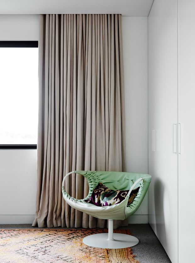 A bright, minimalist interior corner featuring full-height taupe curtains beside a narrow window, white built-in cabinetry with slim handles, and a distinctive pale green sculptural lounge chair with a patterned cushion. A muted patterned rug sits over a dark grey carpet, creating a refined contemporary reading or seating nook.
