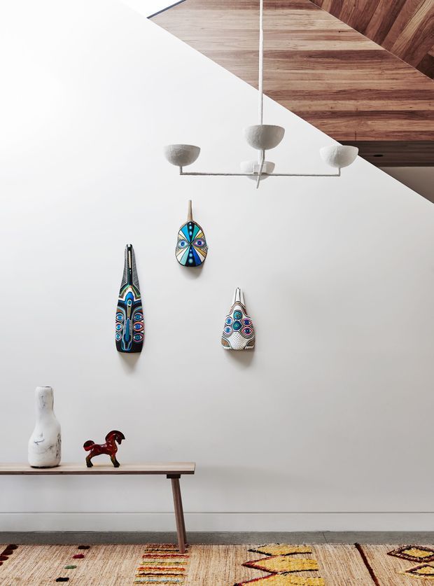 A minimalist interior vignette with a white wall displaying three colourful elongated decorative masks, a slim timber console table with sculptural objects, and a large patterned rug. A simple suspended multi-arm light hangs below a dramatic angled timber-lined ceiling or soffit, adding warm natural texture to the otherwise pale space.