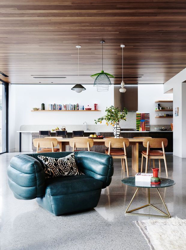 Open-plan contemporary kitchen and living area with a dramatic dark timber-lined ceiling, polished concrete floor, white walls, timber kitchen island and stools, open shelving, sculptural pendant lights, and a distinctive deep teal leather lounge chair with a glass-and-metal coffee table. The space has strong mid-century-inspired furnishings within a modern architectural shell.