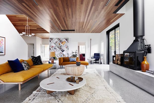 Bright contemporary living room with a mustard-yellow sectional sofa, sculptural round coffee tables, a large textured rug, open geometric shelving, and a substantial black wood-burning fireplace with a stone or concrete hearth. The room features extensive warm timber ceiling lining, white walls, large black-framed windows, and minimalist integrated lighting.