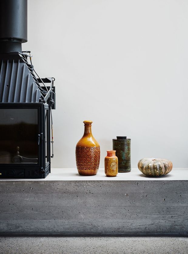A minimalist interior vignette featuring a black cast-iron wood-burning stove with a tall flue on the left, displayed on a raw concrete ledge. Several vintage-style ceramic vessels in amber, orange, and green sit beside a small ribbed decorative object. The setting has a plain light-grey wall, concrete surfaces, and a restrained industrial aesthetic.