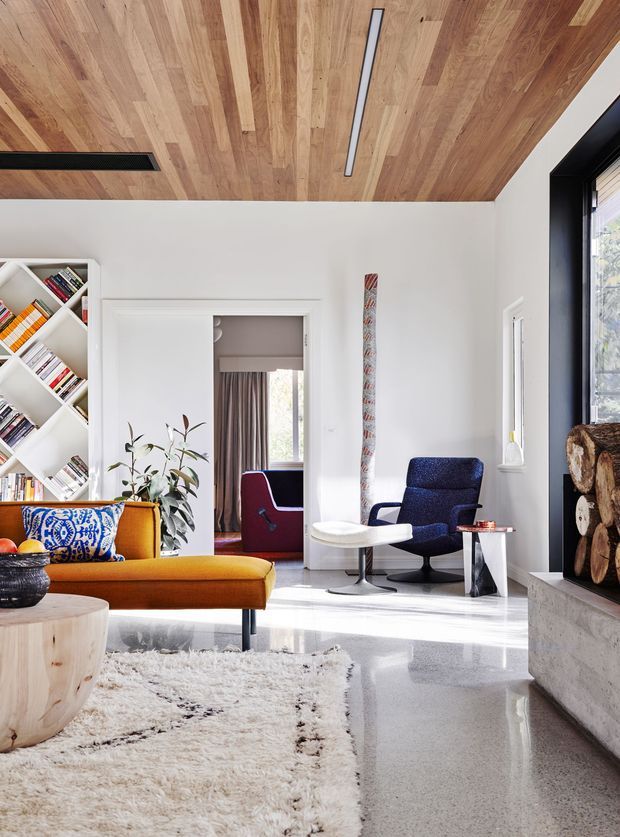 Bright contemporary living room with white walls, a warm timber-lined ceiling, polished concrete flooring, and a black-framed fireplace niche stacked with firewood. The room contains a mustard upholstered sofa, sculptural timber coffee table, blue lounge chair, small side table, patterned cushions, a textured rug, built-in angled bookshelf, indoor plant, and linear recessed lighting. A doorway reveals a curtained adjoining room with another lounge chair.