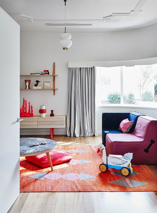A bright, stylish living room viewed through an open doorway, featuring ornate white ceiling mouldings, a sculptural pendant light, pale timber flooring, a colourful geometric rug, a deep blue and burgundy sofa, a low round coffee table, timber wall shelving, and a low timber sideboard. Large bay-window glazing with full-length grey curtains brings in abundant natural light. Children's toys and playful accessories add a family-oriented character.