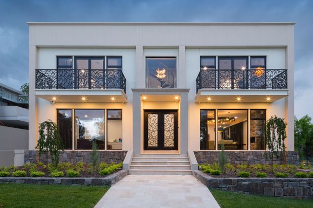 Symmetrical two-storey residential facade photographed from the front at dusk. The pale rendered building has a strong central entry, large dark-framed windows, ornate black metal balcony balustrades, projecting balcony slabs, stone-clad lower walls, landscaped planting beds, and a broad paved approach. Warm exterior lighting and glimpses of a furnished interior are visible through the glazing.