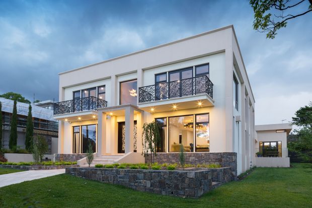 Exterior view of a large two-storey white contemporary house with a clean rectangular form, flat roofline, full-height glazing, projecting balconies with ornate black metal balustrades, warm illuminated interiors, and a dark stacked-stone retaining wall and plinth. The home sits within a landscaped lawn and paved approach.