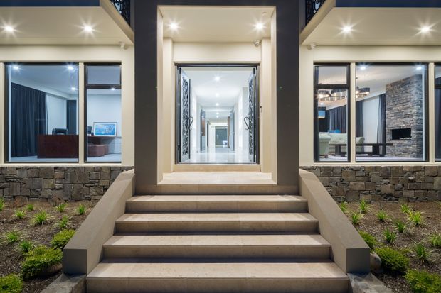 Front-facing exterior view of a contemporary residence at dusk, centred on a broad flight of steps leading to a brightly lit recessed entry. Large windows on either side reveal modern interior spaces, including a living area and a stone-clad fireplace. The facade combines dark vertical elements, light rendered surrounds, extensive glazing, and a low stone-clad base with landscaped planting.