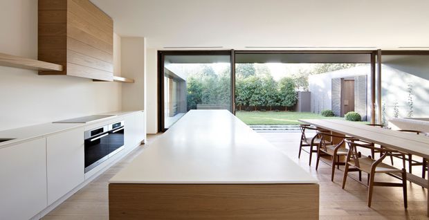 A bright, minimalist open-plan kitchen and dining area. The kitchen features a large white island with a timber-clad base, white integrated cabinetry, timber wall cabinetry and open shelving, and built-in black appliances. Full-height sliding glass doors open to a landscaped courtyard, while a timber dining table and chairs occupy the right side of the room.