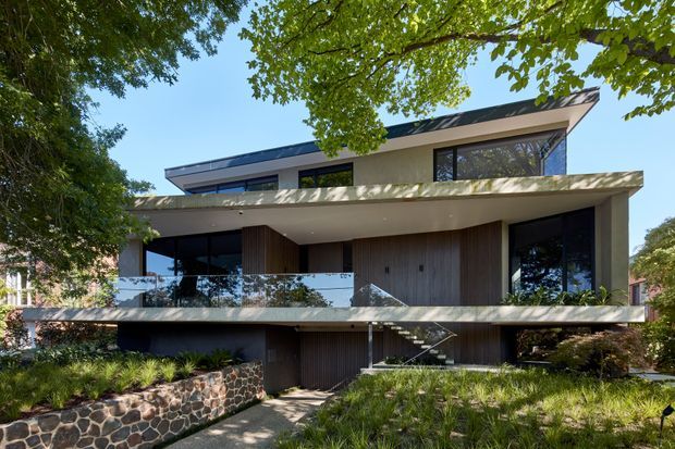 A modern multi-level residence set within a leafy suburban landscape. The facade combines broad horizontal concrete or rendered slabs, extensive dark-framed glazing, vertical timber wall cladding, and a dark low-pitched roof. A glass-balustraded upper terrace and external stair lead through landscaped gardens, with a distinctive rough dark-stone retaining wall in the foreground.