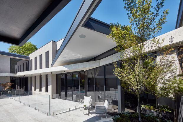 Contemporary multi-wing residence viewed from an outdoor terrace or courtyard. The composition features crisp white rendered walls, dark window and door frames, extensive glazing, strong cantilevered roof planes, and a cantilevered upper-level form. A clear glass balustrade encloses the pale paved terrace, with outdoor dining furniture and landscaped planting around the building. The architecture has a clean, geometric, high-contrast modern character under bright daylight.