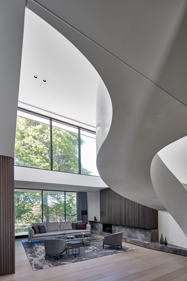 A sophisticated double-height living room with expansive floor-to-ceiling glazing overlooking trees, a large sculptural curved staircase or mezzanine structure, and a contemporary fireplace wall clad in dark vertical timber slats with a stone hearth. The room features a grey sectional sofa, accent chairs, a low glass coffee table, patterned area rug, timber flooring, and restrained neutral furnishings. Bright natural light and strong architectural curves create a refined, dramatic atmosphere.