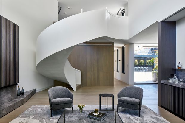 A refined contemporary living room centered on a dramatic sculptural, curved white staircase. The space features pale timber flooring, a large grey patterned rug, two grey upholstered lounge chairs, a low black coffee table, dark vertical-ribbed cabinetry and wall panelling, and a marble-look fireplace or display ledge. Glazing at the rear opens toward an outdoor balcony, while a timber-clad entry or internal door sits beneath the stair.
