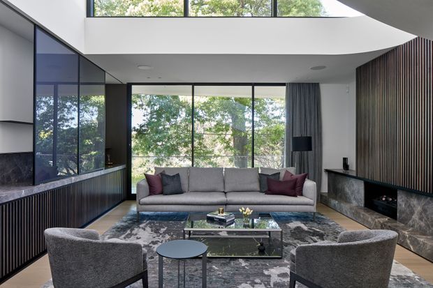 A refined contemporary living room with a large grey sofa, two rounded lounge chairs, a glass coffee table, and a patterned area rug. Full-height glazing and clerestory windows bring in views of trees and abundant natural light. Dark vertical timber-look wall panelling, a dark stone fireplace surround, integrated cabinetry, and black-framed glazing create strong contrast against the white walls and sculptural ceiling opening.