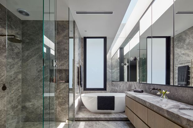 A refined contemporary bathroom featuring a full-height glass shower enclosure, freestanding white bathtub, double vanity with timber-toned cabinetry and a pale veined stone benchtop. Large-format grey stone-look wall and floor surfaces, frameless mirrors, dark window frames, and a linear skylight create a bright, polished interior.