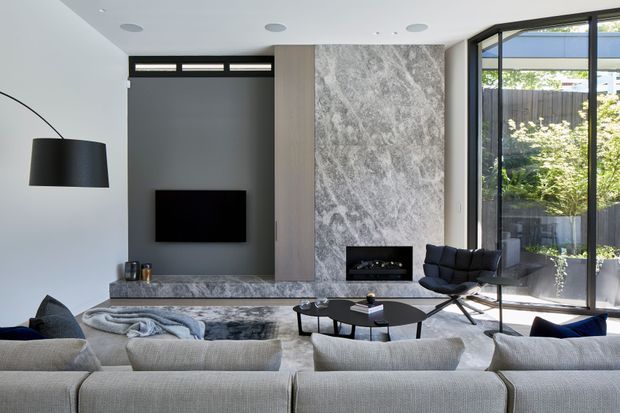 Contemporary living room with a large light-grey sectional sofa in the foreground, black round coffee tables, lounge chairs, and a wall-mounted television. The room features a dramatic grey veined stone feature wall with integrated fireplace, dark timber or charcoal cabinetry, recessed ceiling lights, and full-height glazed doors opening to a planted courtyard.