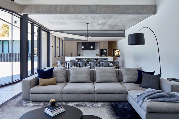 A spacious contemporary open-plan living and dining area with a large grey sectional sofa in the foreground, a round black coffee table, and a dining table with multiple upholstered chairs behind it. Full-height black-framed glazing lines the left side, while a minimalist kitchen with timber-toned cabinetry and a dark island sits at the rear. The room features exposed concrete beams and ceiling, a curved white wall, a large arched floor lamp, layered neutral furnishings, and dark blue accent cushions. The setting appears refined, modern, and open, with an exterior balcony or view visible through the glazing.