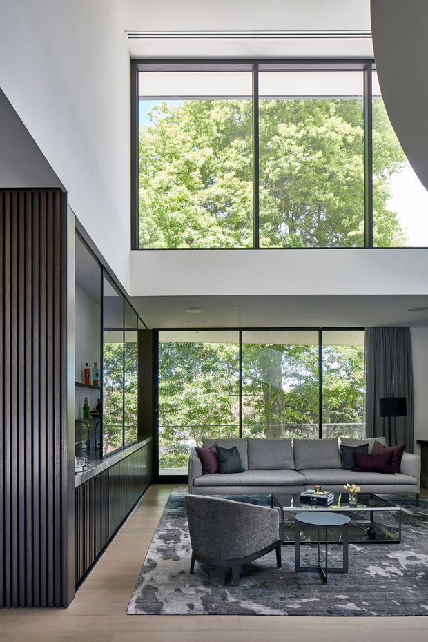 Contemporary double-height living room with expansive black-framed glazing overlooking leafy trees. The room features a large grey sofa, sculptural lounge chairs, a glass coffee table, patterned grey rug, dark timber vertical-slat joinery and an integrated bar or display area. A mezzanine-like upper level and tall windows create a bright, open interior.