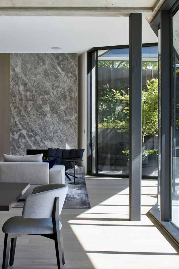 A refined contemporary living and dining area with expansive black-framed glazing opening toward a planted courtyard. The room features a dramatic grey marble or natural-stone feature wall, pale timber flooring, exposed concrete ceiling beams, dark structural columns, a light upholstered sofa, dark lounge chairs, and a dining table with upholstered chairs. Strong sunlight creates long linear shadows across the floor.