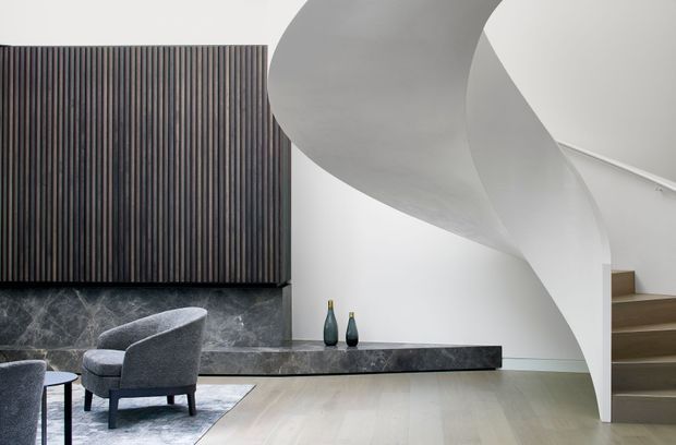Contemporary interior featuring a sculptural curved staircase with a smooth white underside, pale timber flooring, a dark marble fireplace or feature ledge, and a vertical dark timber slat wall. Grey upholstered lounge chairs and a small side table sit at the left, with a minimal decorative arrangement on the ledge.