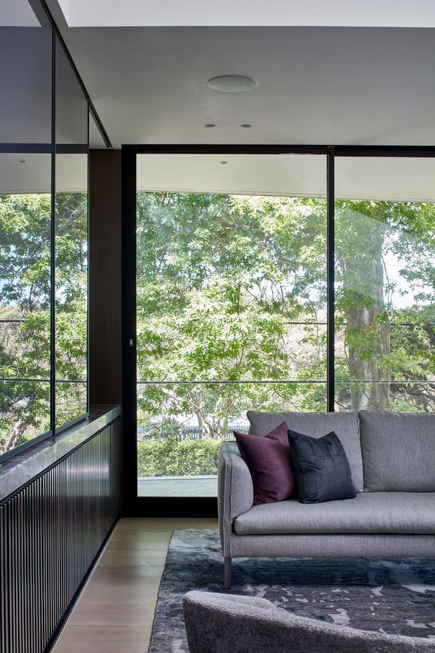 A contemporary living room corner with a grey upholstered sofa, dark accent cushions, and a patterned rug. Full-height black-framed glazing wraps the room, overlooking leafy vegetation. A dark vertically fluted built-in wall or cabinetry element runs along the left side, with light timber flooring and a smooth white ceiling.