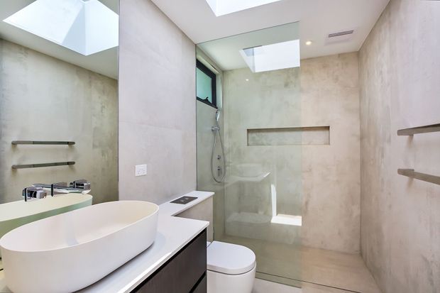 Bright contemporary bathroom with a freestanding white vessel basin on a dark vanity, wall-mounted toilet, frameless glass shower enclosure, recessed wall niche, open shelving, large-format pale stone-look wall and floor finishes, and a skylight providing natural light.