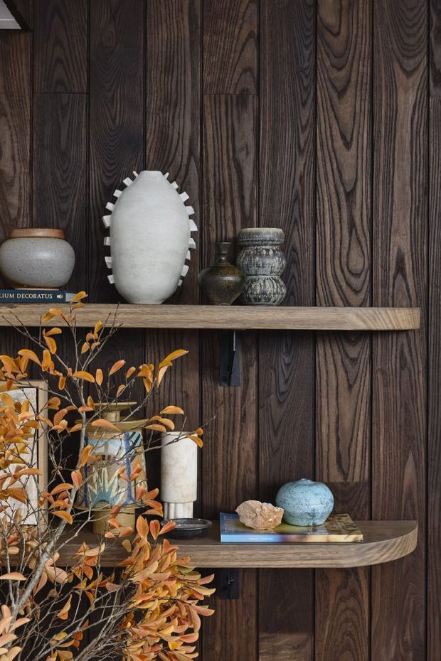 A close-up interior vignette featuring two floating timber shelves mounted against a dark, vertically grained timber-panelled wall. The shelves display sculptural ceramic and stone vessels, books, and decorative objects, with dried orange-toned foliage in the foreground. The composition is richly textured, warm, and moody, with strong natural-material detailing and curved shelf edges.