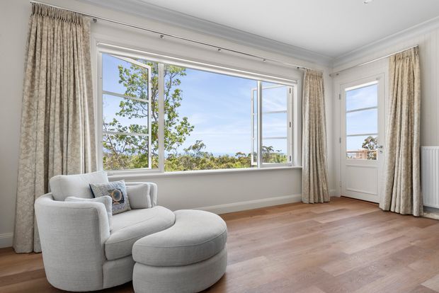 Bright, refined sitting room with a large bay-style window and glazed exterior door, framed by full-length textured curtains. The room features a pale upholstered armchair with matching ottoman, white walls with decorative cornice detailing, timber flooring, and views of leafy surroundings outside.