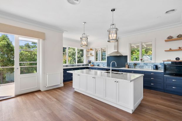 Bright, spacious kitchen with a large white island, navy-blue perimeter cabinetry, pale stone benchtops, a blue patterned tiled splashback, pendant lighting, timber flooring, and white-framed windows and glazed doors opening toward a leafy exterior.