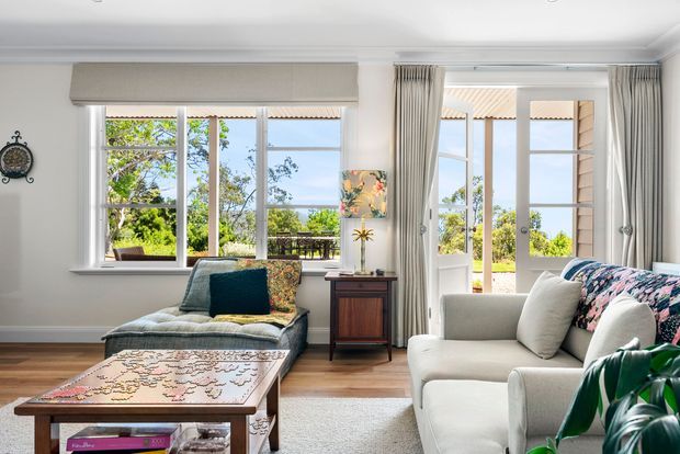 Bright, relaxed living room with white walls, timber flooring, large windows and glazed doors opening to an outdoor terrace. The room includes a cream sofa, upholstered chaise lounge, patterned coffee table, curtains, a timber side cabinet and leafy greenery, with abundant natural light and views of greenery and water beyond.