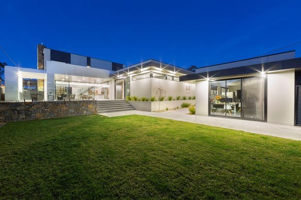 Twilight exterior view of a low, contemporary flat-roofed residence arranged around a landscaped lawn and paved courtyard. The home features crisp white and dark charcoal facade planes, extensive floor-to-ceiling glazing, illuminated covered outdoor areas, broad steps, and a textured stone retaining wall. Interior dining and living spaces are visible through the glass.